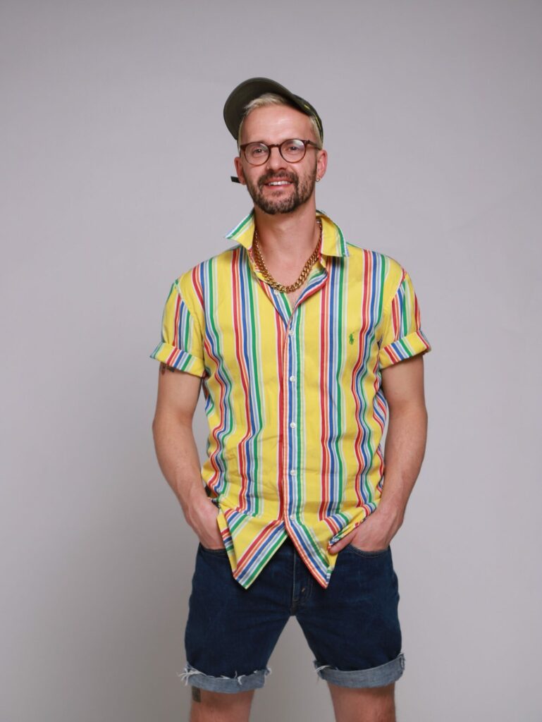 a photo of the author Dan Glass standing against a grey background, striped shirt and denim shorts
