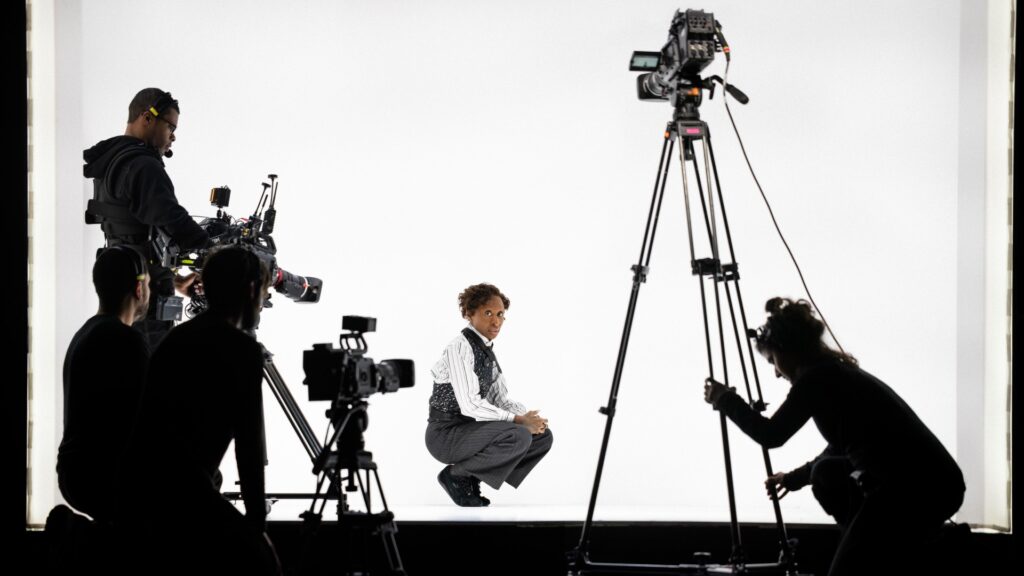 Cynthia Erivo on stage in Dracula at No&euml;l Coward Theatre in white shirt and black trousers, against a brightly lit white backdrop and with cameras around her