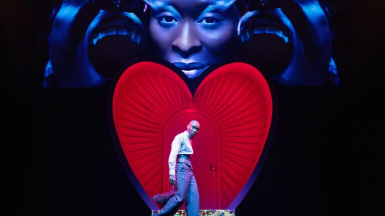 Cynthia Erivo on stage in Dracula at Noël Coward Theatre in white shirt and grey trousers with a red heart behind her (Image: Daniel Boyd)