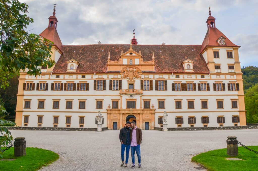 Two men hold hands in front of the baroque Eggenberg Palace