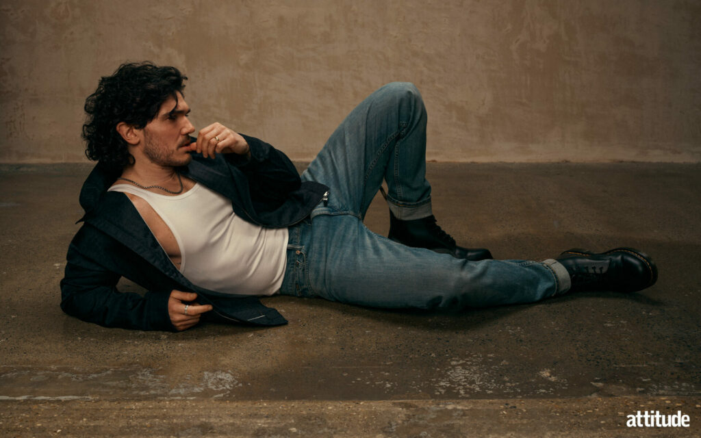 Fra Fee lying on the floor with one leg bent, wearing a white vest and an open black hoodie paired with blue jeans and black Doc Marten boots