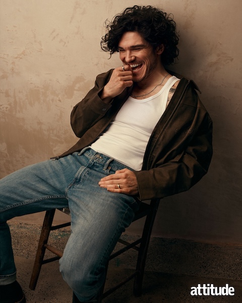 Fra Fee, wearing a white vest, brown cardigan and blue jeans, sat on a chair biting his hand whilst smiling 