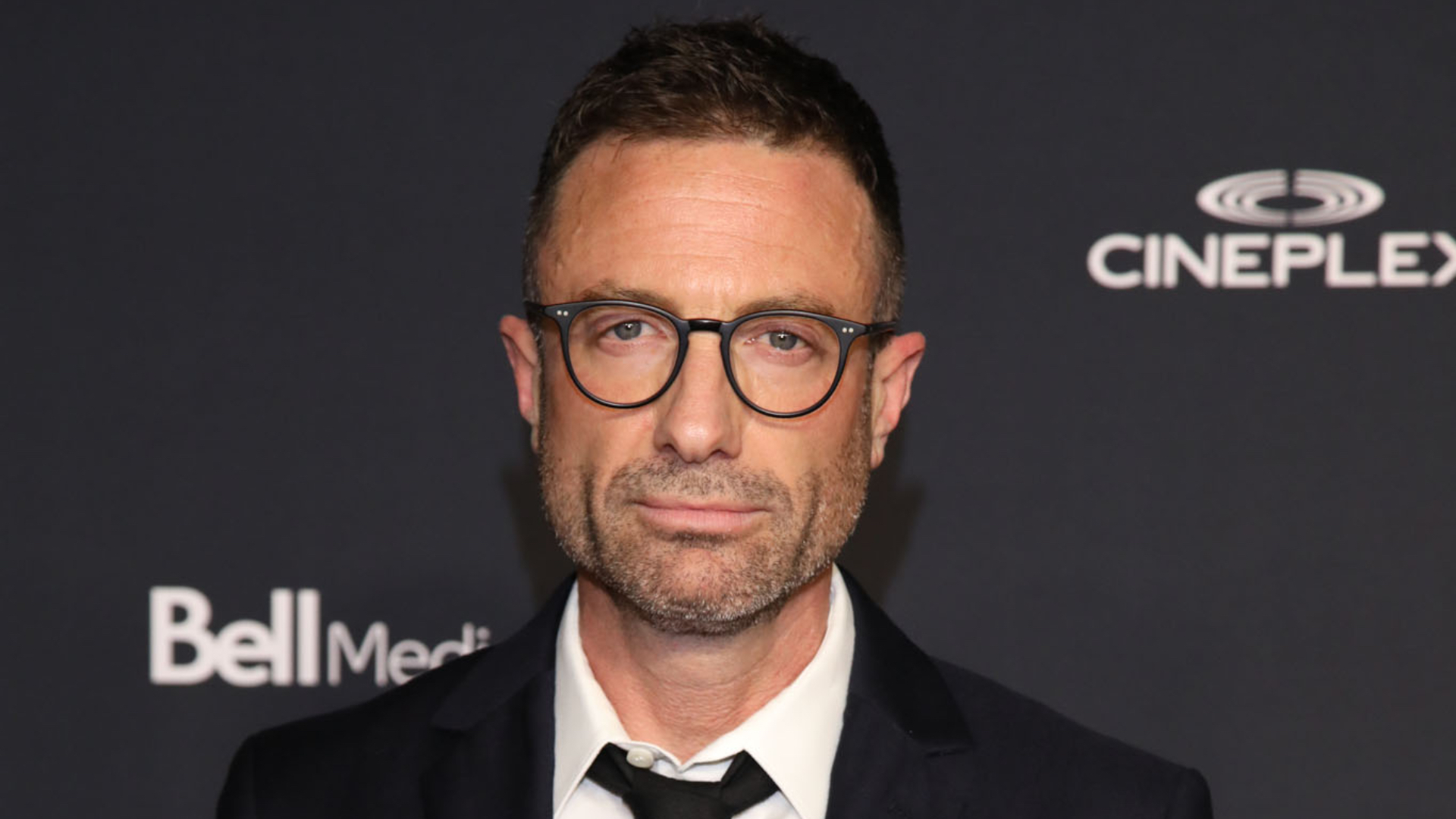 Jacob Tierney in a white shirt and black tie under a black blazer, paired with glasses, on the red carpet