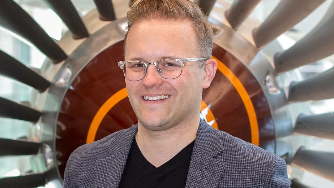 A smiling man in glasses and a gray blazer standing in front of a jet engine