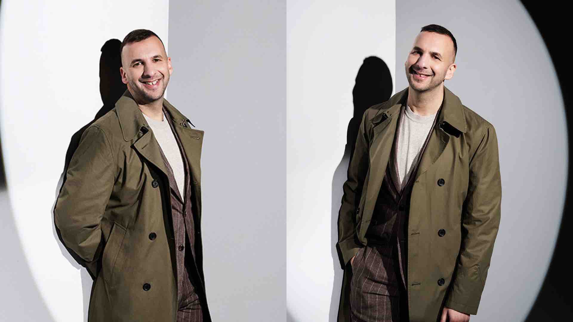 Zack Polanski stands in a green trench coat, smiling as a spotlight lights him up through the darkness