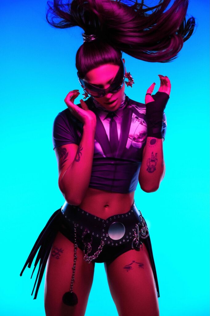 Pabllo Vittar, under pink lighting with their ponytail flying through the air, wore sunglasses, a cropped T-shirt, a large belt, and shorts