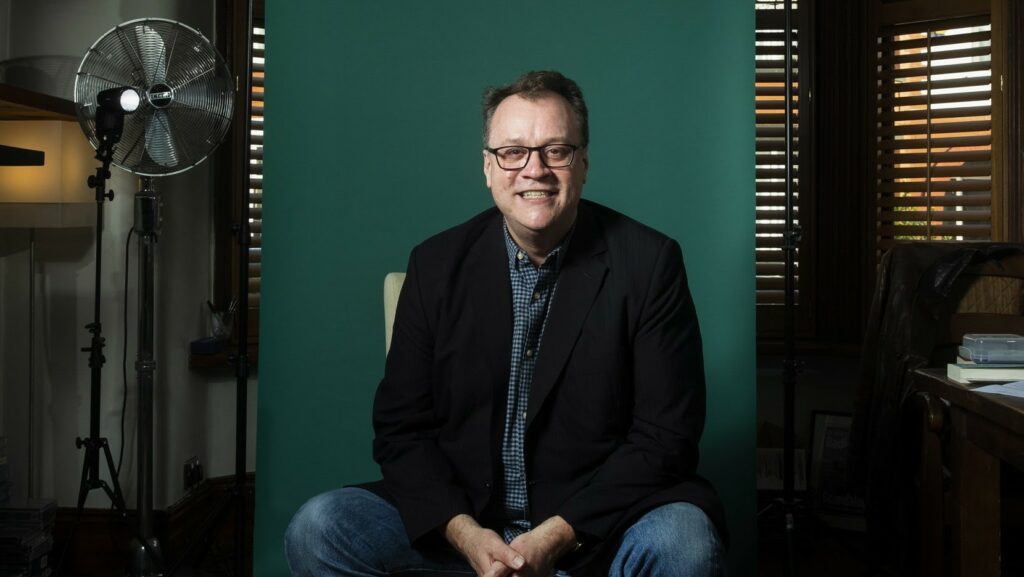 Russell T Davies in a black suit jacket and checked blue shirt, smiling on a chair with his hands together