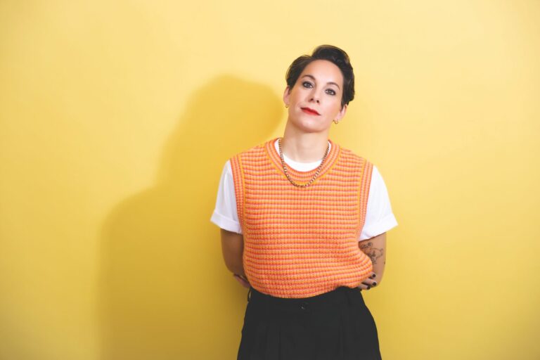 Suzi Ruffell wearing an orange jumper and black trousers standing against a yellow backdrop
