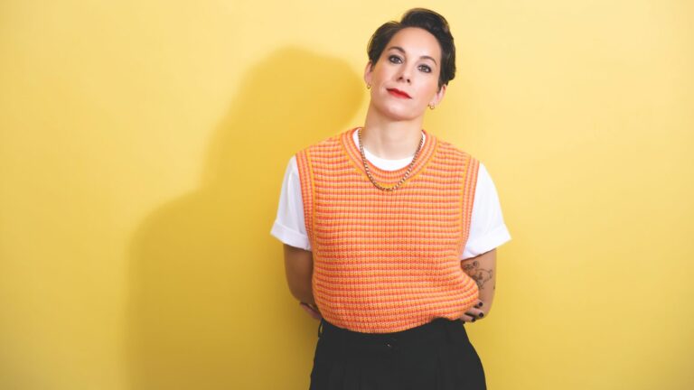 Suzi Ruffell wearing an orange jumper and black trousers standing against a yellow backdrop