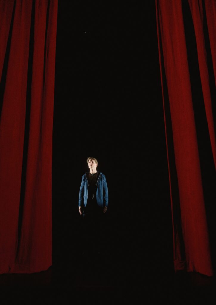 Sean Hayes in The Unknown dimly lit by an overhead light centred in the middle of two red curtains