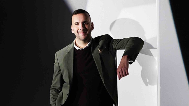 Zack Polanski wearing a green blazer over a black jumper, leaning casually against a white podium