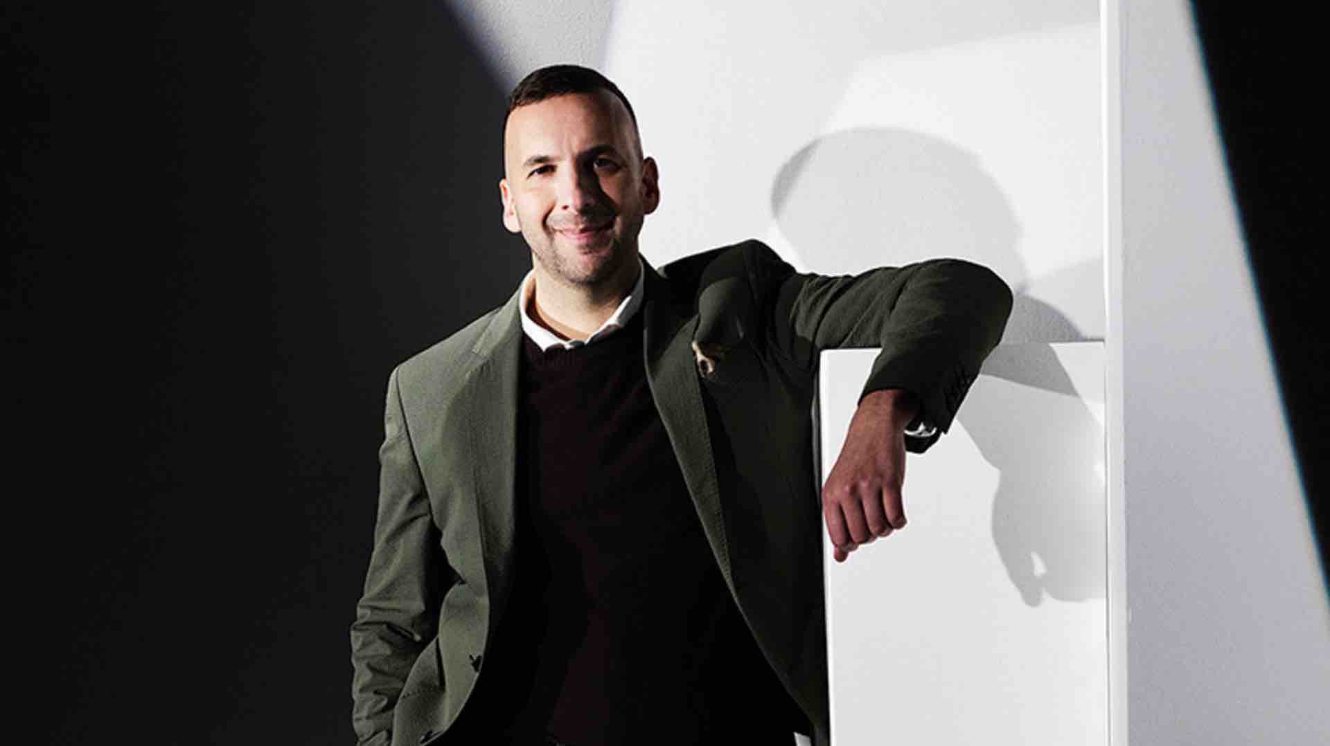 Zack Polanski wearing a green blazer over a black jumper, leaning casually against a white podium