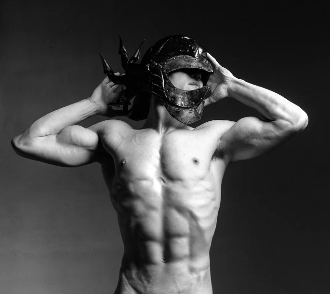 Black-and-white portrait of a shirtless man wearing a spiked metal helmet, holding it with both hands against a dark background.