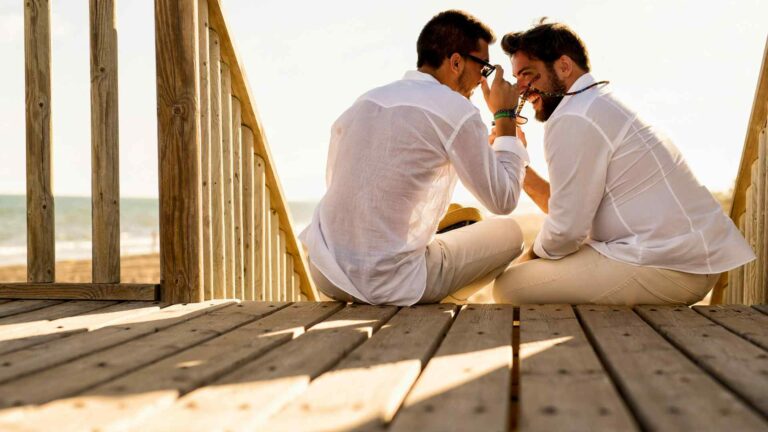 Two men in summery outfits sitting on a deck by a beach