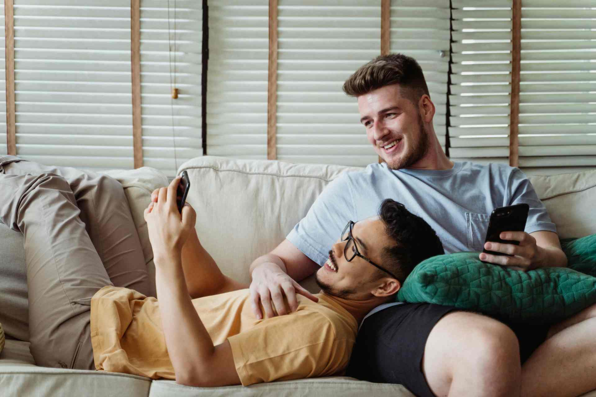 Two men lying on a sofa smiling and using their phones