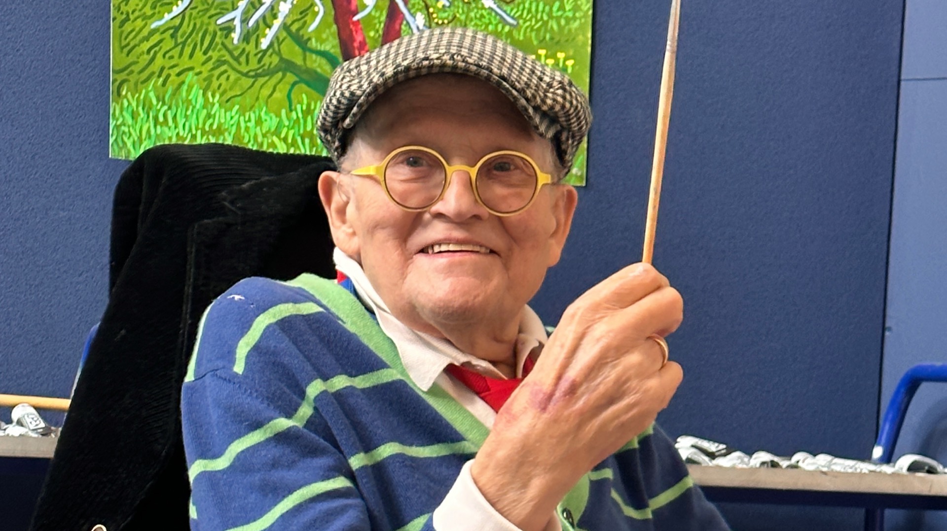 David Hockney photographed in London in 2023, holding a paintbrush and wearing a hat and blue striped top (Image: Supplied)