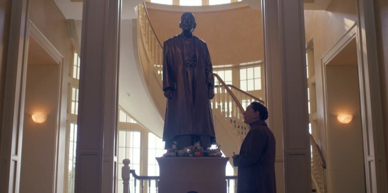 A still from The Testaments showing Aunt Lydia looking at a statue of herself in front of a winding staircase