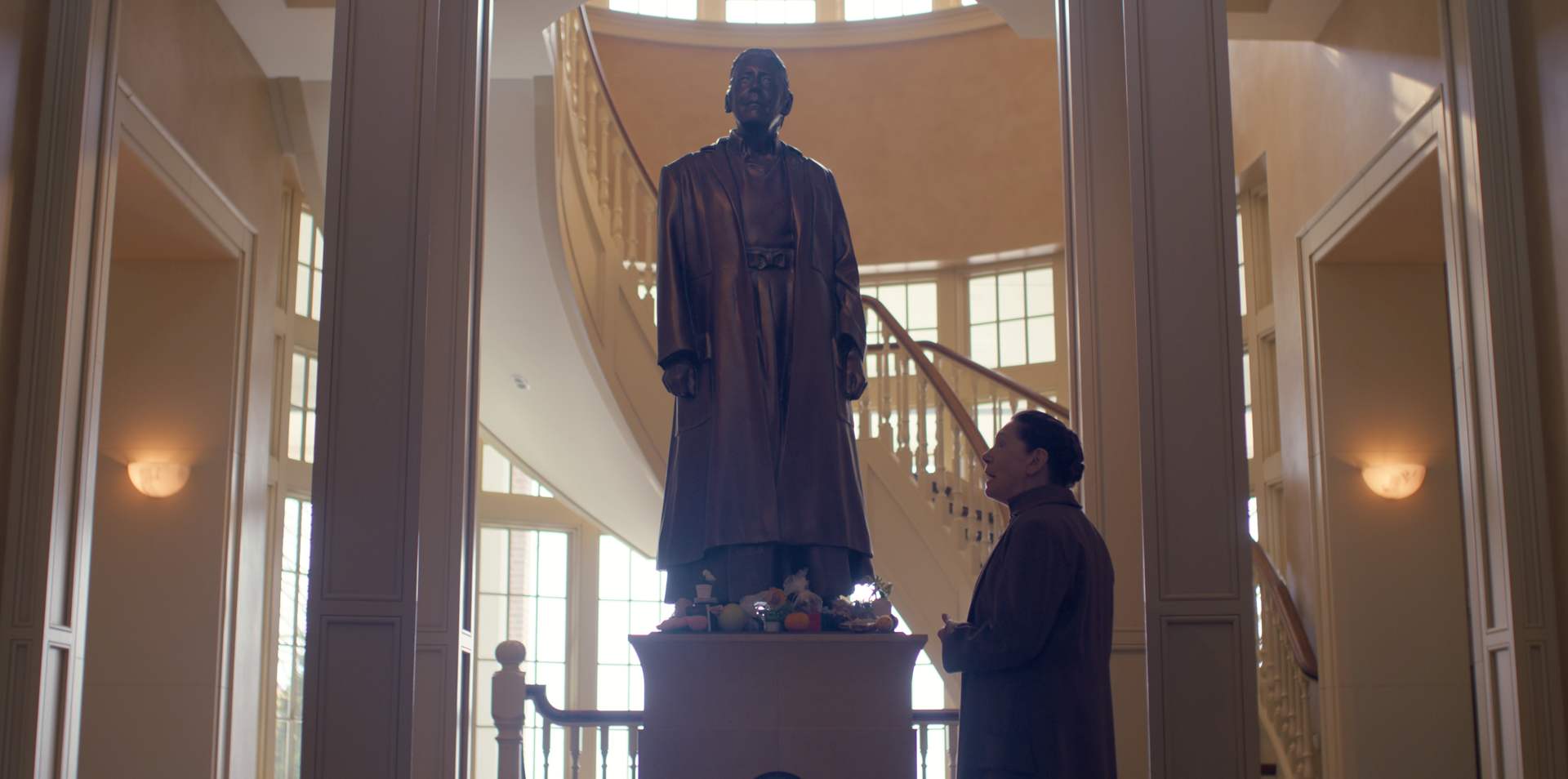 A still from The Testaments showing Aunt Lydia looking at a statue of herself in front of a winding staircase
