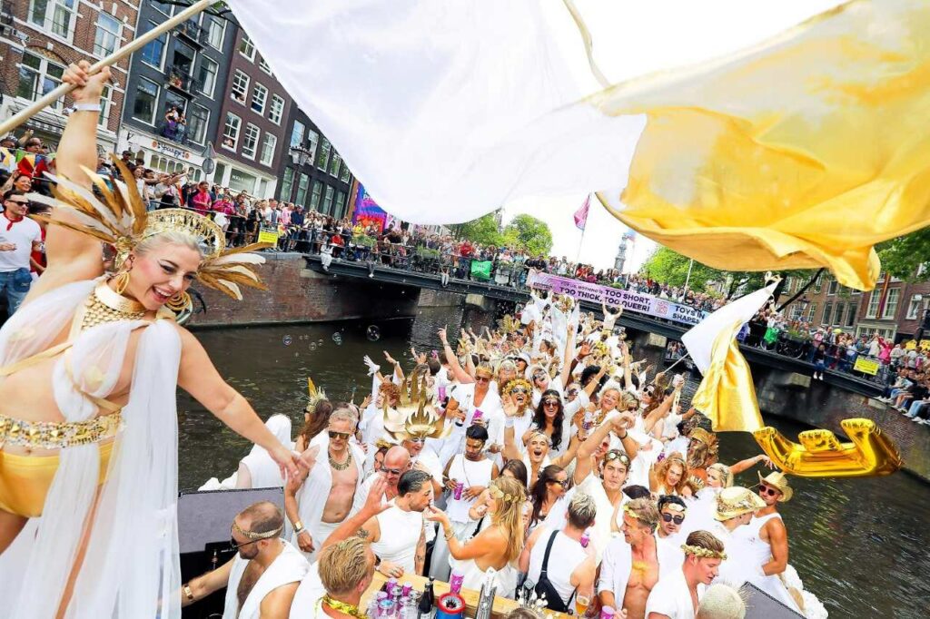 A large group of people dressed in white celebrate on a canal boat during Amsterdam Pride, with crowds of spectators watching from the banks and a bridge in the background. In the foreground, a person wearing an elaborate gold headdress and white outfit holds up a large white and yellow flag.