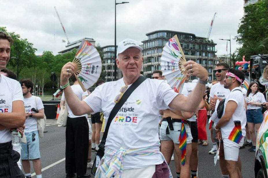 Actor Paul Clayton holding up two Pride-themed hand fans while marching in an Attitude t-shirt at the 2026 Pride in London parade.