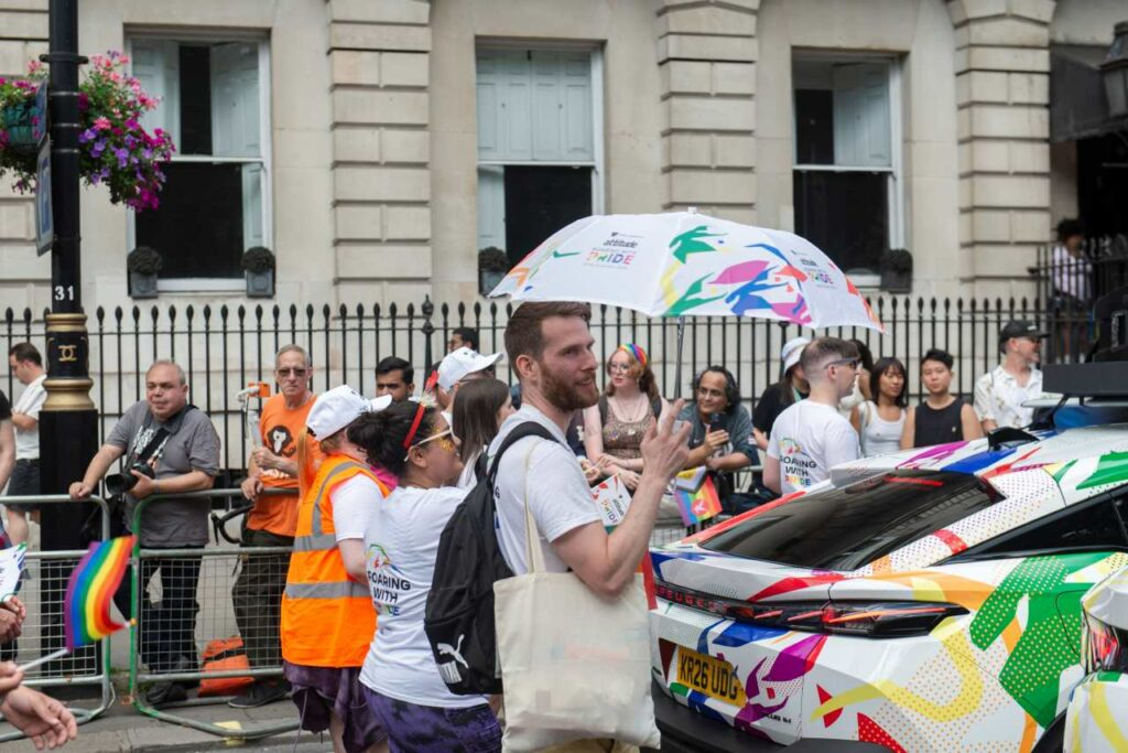 A participant holding an Attitude Pride umbrella, standing near a Peugeot with a colourful wrap at the 2026 Pride in London parade