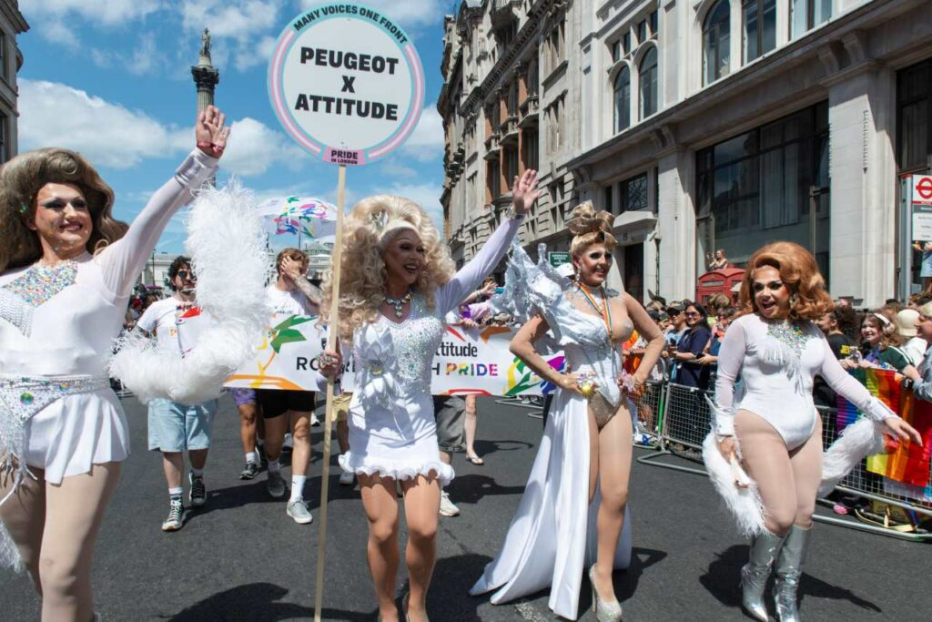 Miss Tess Tickle & The Dragettes in white and silver outfits marching at Pride in London, holding a Peugeot x Attitude sign.