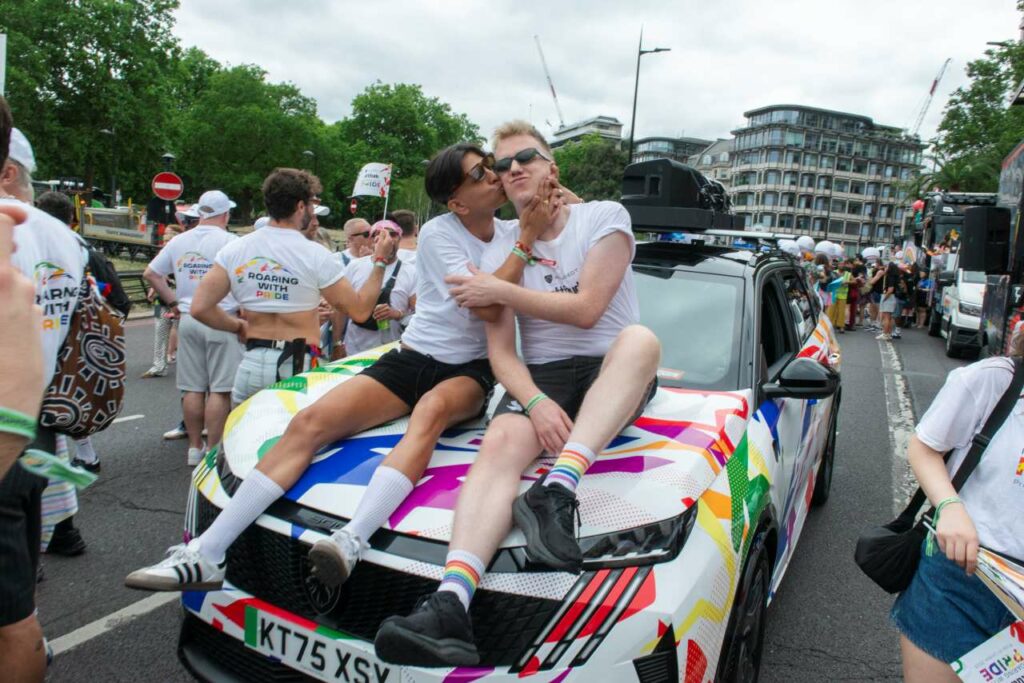 Jason Patel and James Barr sitting on the bonnet of a Pride-wrapped Peugeot car at the 2026 Pride in London parade, with Jason kissing James on the cheek.