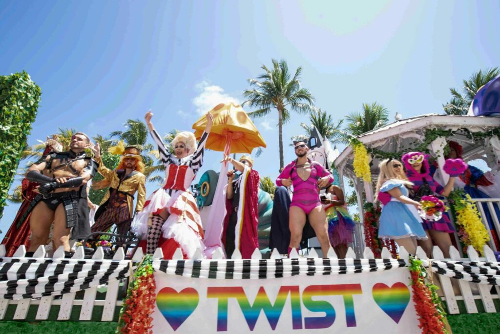 A group of performers in costume pose on the Twist float at the 2025 Miami Beach Pride, with a sign in front featuring the word "TWIST" flanked by rainbow hearts.