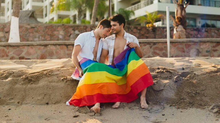 Two men sat on a beach holding a Pride flag