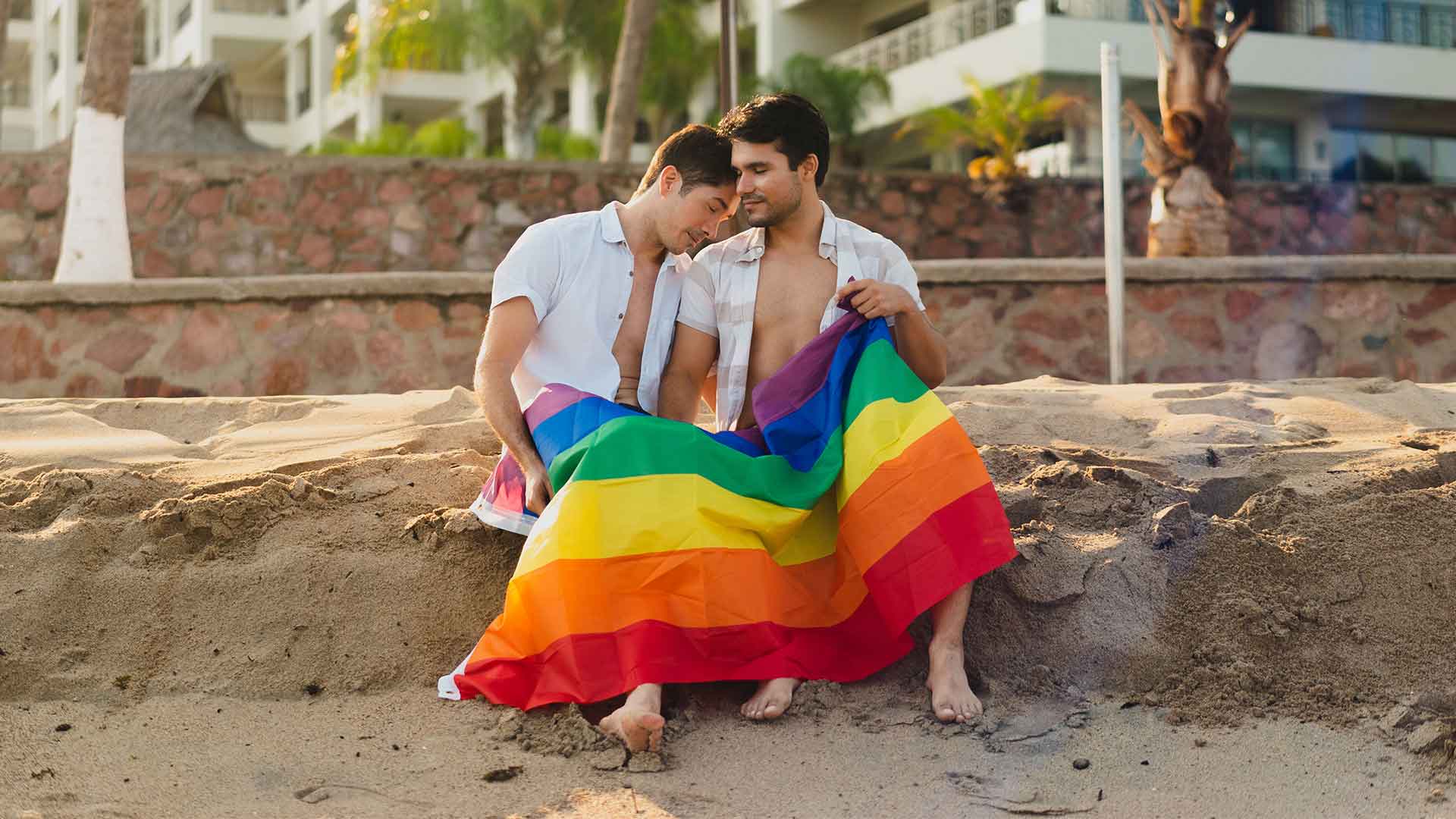 Two men sat on a beach holding a Pride flag