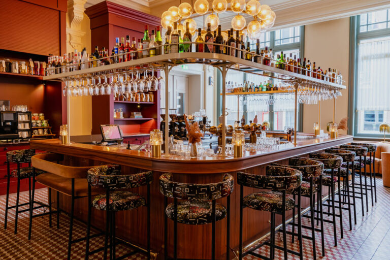 A large four counter island bar with bar stools all around it and a grand chandelier above it.