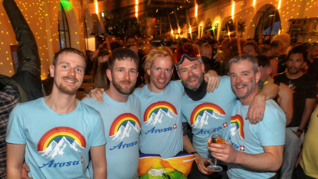 A group of five smiling men wearing matching light blue Arosa mountain t-shirts pose together at an après-ski party during Arosa Gay Ski Week.