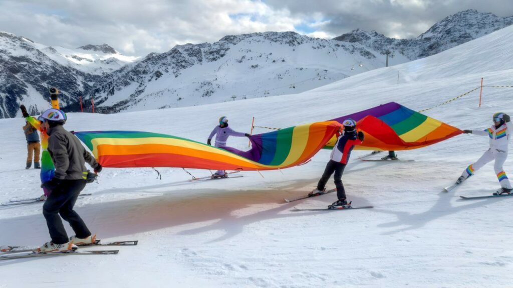 Skiers holding a giant rainbow pride flag across a snowy slope during Arosa Gay Ski Week.