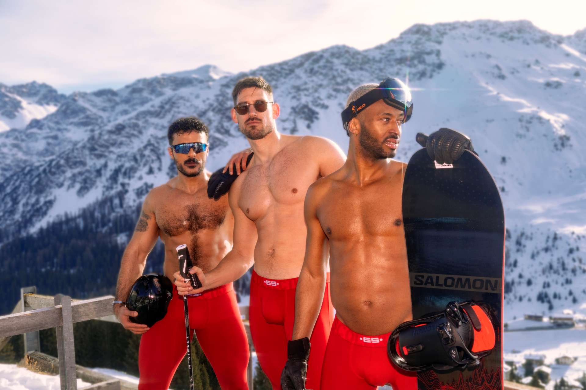 Three shirtless men pose in red ski trousers with a snowboard on a snowy mountain during Arosa Gay Ski Week.