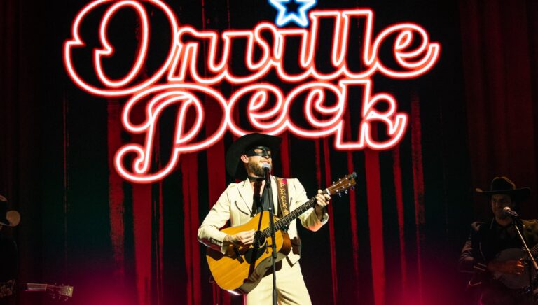 Orville peck in white jacket, eye mask, holding a guitar and standing before a neon Orville Peck sign