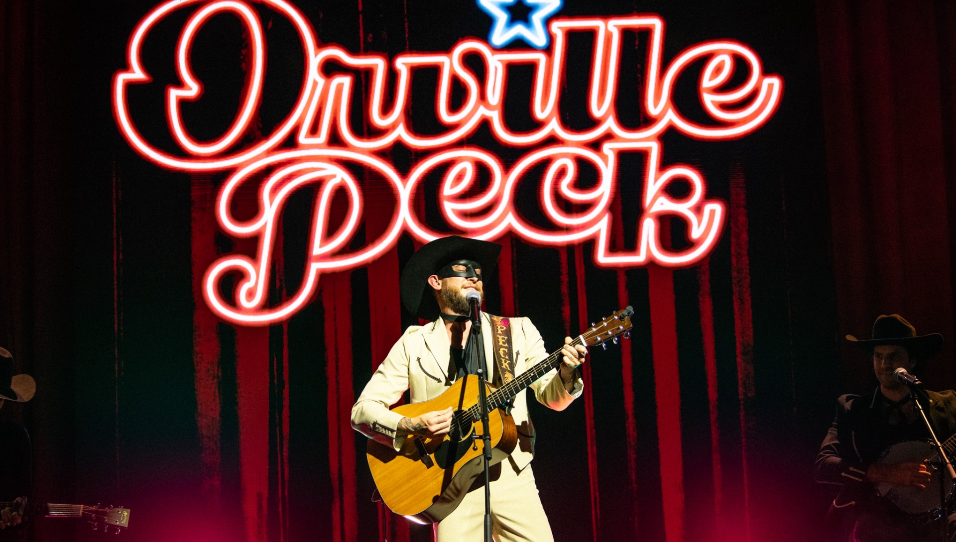 Orville peck in white jacket, eye mask, holding a guitar and standing before a neon Orville Peck sign